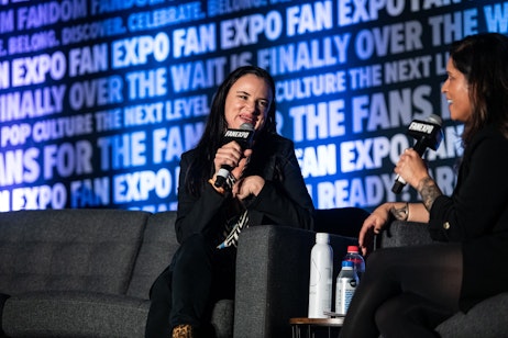 The image shows Juliette Lewis speaking into a microphone, likely a panelist at a FAN EXPO event, as indicated by the backdrop. She has a joyful expression, perhaps sharing an amusing story or answering a question, and appears to be mid-sentence. Her relaxed posture and engaging smile suggest a comfortable and interactive atmosphere, common in panel discussions or Q&A sessions at fan conventions. The backdrop emphasizes the theme of the event, celebrating fandom and pop culture.