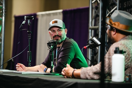 The image shows comic artist Ryan Ottley, wearing a casual gray and green baseball cap and a dark shirt. He has a friendly and relaxed demeanor, with a slight smile as he looks off to the side, possibly engaging with someone or listening attentively. Microphones on the table suggest he is part of a discussion or interview at a convention or public event. The setting has a professional atmosphere with stage equipment in the background, and the presence of other panelists or attendees can be inferred from the cropped figures in the foreground. His smartwatch and casual attire lend a contemporary and approachable vibe to his appearance.