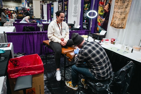 The image depicts a tattoo artist at work on a client's leg. The client, a woman, is seated comfortably on a bench, looking down at the artist with a pleased expression. She's wearing a casual white sweatshirt, leggings, and sneakers. The tattoo artist, wearing a grey plaid shirt and a beanie, is focusing intently on the tattooing process, with his gloved hands applying the tattoo on the woman's leg. In the background, there's a purple curtain that partitions their space, and various artwork and supplies are visible, suggesting this is at a tattoo convention or a pop-up booth. There is a ring light providing ample lighting for the artist to work by. The overall setting seems relaxed and informal, with a black protective sheet covering the floor, indicating care for cleanliness.