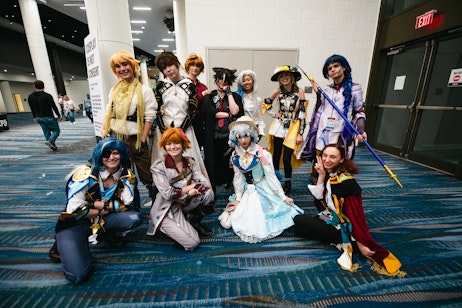 The image is a lively group photo of cosplayers at a convention. Each individual is dressed in elaborate costumes, likely representing characters from anime, video games, or other pop culture media. Their outfits feature a variety of colors, patterns, and accessories such as wigs, hats, and weapons. The cosplayers are posing with enthusiasm and confidence, some standing while others kneel or sit, showcasing the details of their costumes. The background is an indoor convention space. The group's attire and the convention setting suggest a shared enthusiasm for costume design and character portrayal.