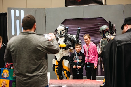 The image depicts a scene from a fan convention where two children are posing for a photo with a person in elaborate cosplay armor, resembling characters from a popular science fiction universe. The cosplayer is wearing a helmet and armor, with details suggesting a high level of craftsmanship and attention to the original character design. One child seems particularly thrilled, standing close to the cosplayer, while the other appears more reserved. An onlooker is taking a photo, capturing this memorable moment. The backdrop gives the impression of a themed booth or photo area, enhancing the immersive experience of the event. The kids' expressions and the overall setup suggest that this is a special moment for young fans meeting their fictional heroes come to life.