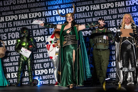 The image shows a person in a vibrant green and gold cosplay costume, complete with a horned helmet, striking a triumphant pose at what appears to be a costume contest or cosplay event. Behind them, individuals in various other costumes add to the festive atmosphere. The backdrop features the "FAN EXPO" logo, indicating the setting is a fan convention. The cosplayer's open arms and wide smile convey a sense of celebration and enthusiasm, embodying the spirit of cosplay where fans creatively express their favorite characters.