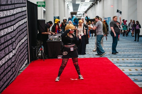 The image shows a person striking a playful pose on a red carpet, likely cosplaying a character from popular media. They are wearing a black outfit with distinctive accessories, including a large belt and patterned tights, paired with sneakers. Their hair is styled in a blonde bob with bangs, and they're holding their hands up in fists, possibly as part of their character's persona. In the background, there's a DJ booth with someone operating equipment, suggesting this is an event with music and entertainment, such as a convention or fan expo. Other people in the background are also dressed in various costumes, indicating a cosplay-friendly environment. The atmosphere is energetic, with the central figure engaging with the camera and the crowd, adding a dynamic element to the scene.