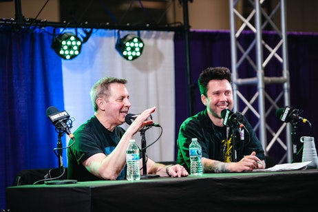The image captures two men seated at a panel, both smiling and engaged in a discussion. The man on the left is gesturing with his hand, likely making a point, or telling a story, while the man on the right listens with a cheerful expression. They are both dressed casually and have microphones in front of them, indicating they are part of a conversation or interview at an event. The backdrop is not fully visible, but the setting appears to be a stage with professional lighting, typical of conventions or expos. Their engaging interaction and the water bottles on the table suggest a relaxed yet organized atmosphere.
