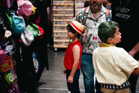 The image features a young child wearing a straw hat and a red vest, looking with interest at a display of colorful backpacks and plush toys at what appears to be a convention or market. To the right, part of an adult, presumably accompanying the child, is visible. The variety of merchandise and the child's intrigued expression capture the wonder and excitement often seen in children when they are exploring and discovering new things they enjoy. The setting suggests an environment where pop culture merchandise is sold, such as a comic book or anime convention.