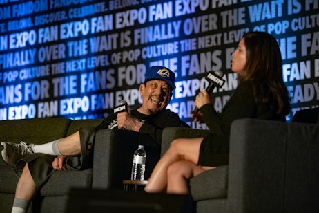 The image shows a lively interaction at a FAN EXPO panel discussion. Danny Trejo, wearing a cap, is laughing expressively, possibly in response to a question or comment. A woman, likely the moderator or interviewer, is holding a microphone and appears to be asking a question or engaging in conversation with him. The man's animated expression suggests a humorous or light-hearted moment. They are both seated in an event setting with a backdrop that reads "FAN EXPO," indicative of a fan convention or cultural event. The focus of the image is on their interaction, capturing a moment of genuine amusement and engagement.