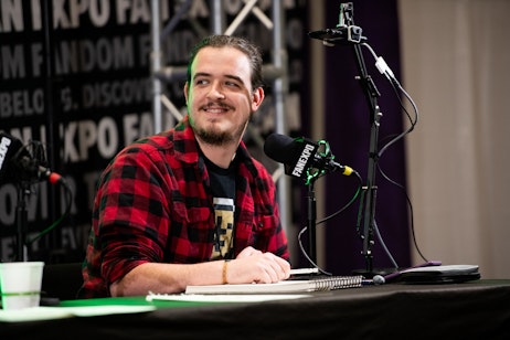 The image features comic artist Gavin Guidry wearing a red and black plaid shirt, sitting at a table with a microphone, appearing to be part of a panel. He has a content and thoughtful expression, looking slightly away from the camera, with a hint of a smile. He's wearing a bracelet on his wrist and has a casual, approachable demeanor. The backdrop, prominently displaying "FAN EXPO" logos, suggests the setting is a fan convention or similar pop culture event. The green stage lighting casts a glow on him, highlighting his presence as a speaker or panelist at the event. His relaxed posture and the notebook on the table in front of him indicate he might be preparing to speak, take notes, or engage in a discussion.