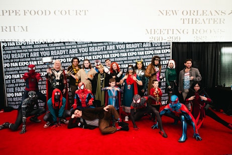 The image is a group photo of individuals in various cosplay outfits, likely taken at a fan expo event. The group includes characters that resemble superheroes and figures from popular culture, showcasing a diverse array of costumes with vibrant colors and designs. Some individuals are posing heroically, while others are in playful stances, contributing to the dynamic and festive atmosphere typical of comic conventions and cosplay gatherings. The backdrop with "FAN EXPO" and "FOOD COURT" text indicates the event location and setting. The overall scene is one of creativity, enthusiasm, and community spirit among fans of pop culture.