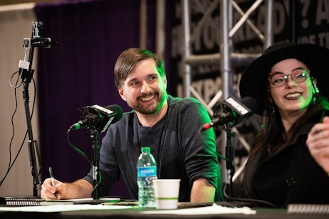 The image shows Comic Creator Travis Hymel and a female moderator sharing a light-hearted moment, both smiling and seemingly enjoying a conversation. They are seated at a table with microphones, suggesting they are part of a panel at an event. Travis has a beard and is wearing a casual shirt, while the woman is stylish with a black hat and glasses, and what appears to be a smile reflecting in the green light from the stage. The backdrop with the words "FAN EXPO" implies they are at a fan convention or similar event, engaging with an audience or participating in a discussion related to pop culture. The atmosphere is relaxed and jovial, indicating a friendly and interactive session.