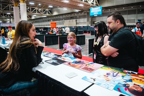 The image shows an interaction at a convention booth, where a young girl, smiling and wearing glasses, is standing across from Sarah Natochenny. The girl seems to be holding an item, possibly for an autograph or discussion, and Sarah appears to be in conversation with her, with a thoughtful expression. In the background, another individual in cosplay and signs indicating "CELEBRITY AUTOGRAPH" area can be seen, denoting the setting as a fan expo or similar event where attendees meet with creators, artists, or celebrities. The layout of the venue, with its spacious interior and designated areas, suggests a large convention center. The atmosphere is engaging and friendly, characteristic of these fan gatherings.