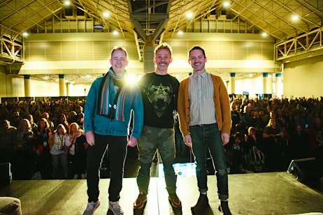 The image captures a joyful moment with Billy Boyd, Dominic Monaghan, and Elijah Wood standing on a stage, smiling and posing for the audience. They are backlit by a bright light, suggesting they are at a significant event or conference. The crowd in the background is blurry but appears to be a large, attentive audience, some of whom are holding up phones to capture the scene. The men are dressed casually but with distinct personal styles, indicating they may be special guests or speakers at the event. The atmosphere is vibrant and lively, reflective of a positive interaction between the speakers and their audience.