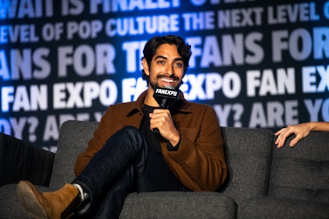 The image shows actor Eman Esfandi seated comfortably with a microphone in hand, mid-conversation. He has a warm smile and is sporting a well-groomed beard, looking attentive and engaged. He's dressed in a dark t-shirt and a brown blazer, giving off a casual yet polished vibe. The backdrop, emblazoned with "FAN EXPO" and phrases about pop culture, indicates that he is likely a guest speaker at a fan or comic convention. His relaxed posture and the intimacy of the setting suggest an interactive session, possibly a Q&A, where he is sharing insights or anecdotes with an audience.