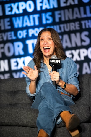 The photo shows Diana Lee Inosanto with an open, joyful expression, speaking into a microphone at what appears to be a panel discussion. She's wearing a denim shirt and has a smartwatch on her wrist. The backdrop features the "FAN EXPO" logo, suggesting she is at a pop culture convention, likely sharing a story or answering questions. Her animated hand gestures and expressive face indicate an engaging and lively conversation.
