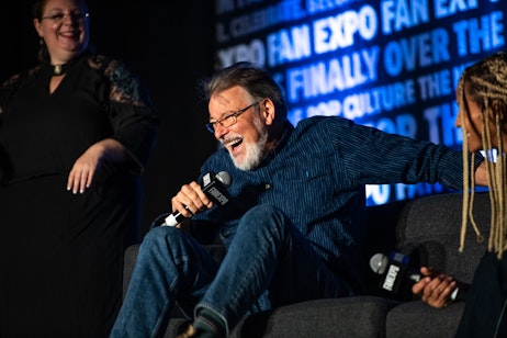 The image features Jonathan Frakes with a microphone, laughing heartily while seated, giving an impression of joy and entertainment. He is casually dressed in a blue shirt and jeans, suggesting a relaxed setting. The backdrop with "FAN EXPO" text indicates that he is likely at a fan convention or similar event, possibly participating in a panel discussion or a public interview. His engaging expression and body language convey that he is enjoying the moment, contributing to a lively and friendly atmosphere.