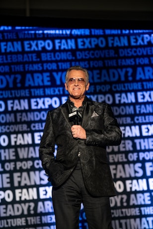The image features actor Bruce Campbell standing confidently in front of a microphone, addressing an audience. He's wearing a dark, patterned blazer with a stylish shirt underneath and sunglasses, suggesting he has a cool and collected persona. The backdrop is emblazoned with the words "FAN EXPO" and other text related to pop culture, indicating that he may be a speaker or host at a fan convention. His demeanor, attire, and handheld microphone all suggest he's likely a key figure at the event, possibly introducing a segment or making an announcement. The lighting focuses on him, making him the clear subject of the photo and likely the center of attention at the event.