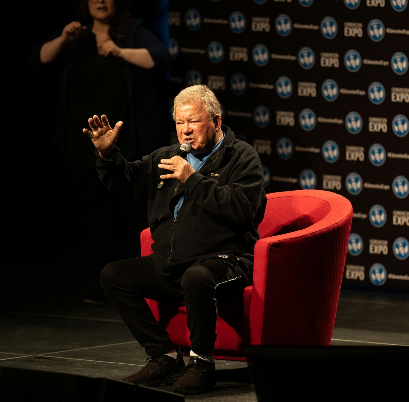 William Shatner sitting in red chair and talking into a microphone.