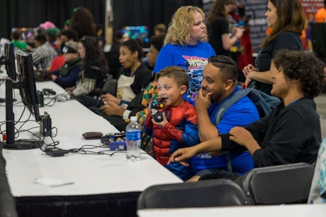 A little boy playing with a controller (in his mouth), while his dad plays with him