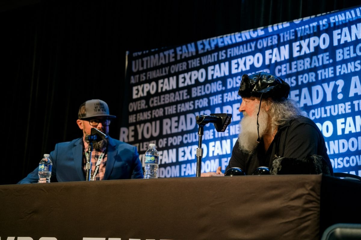 RANDY QUAID AS COUSIN EDDIE SITTING AT A TABLE WITH A MICROPHONE WITH A MODERATOR SITTING NEXT TO HIM