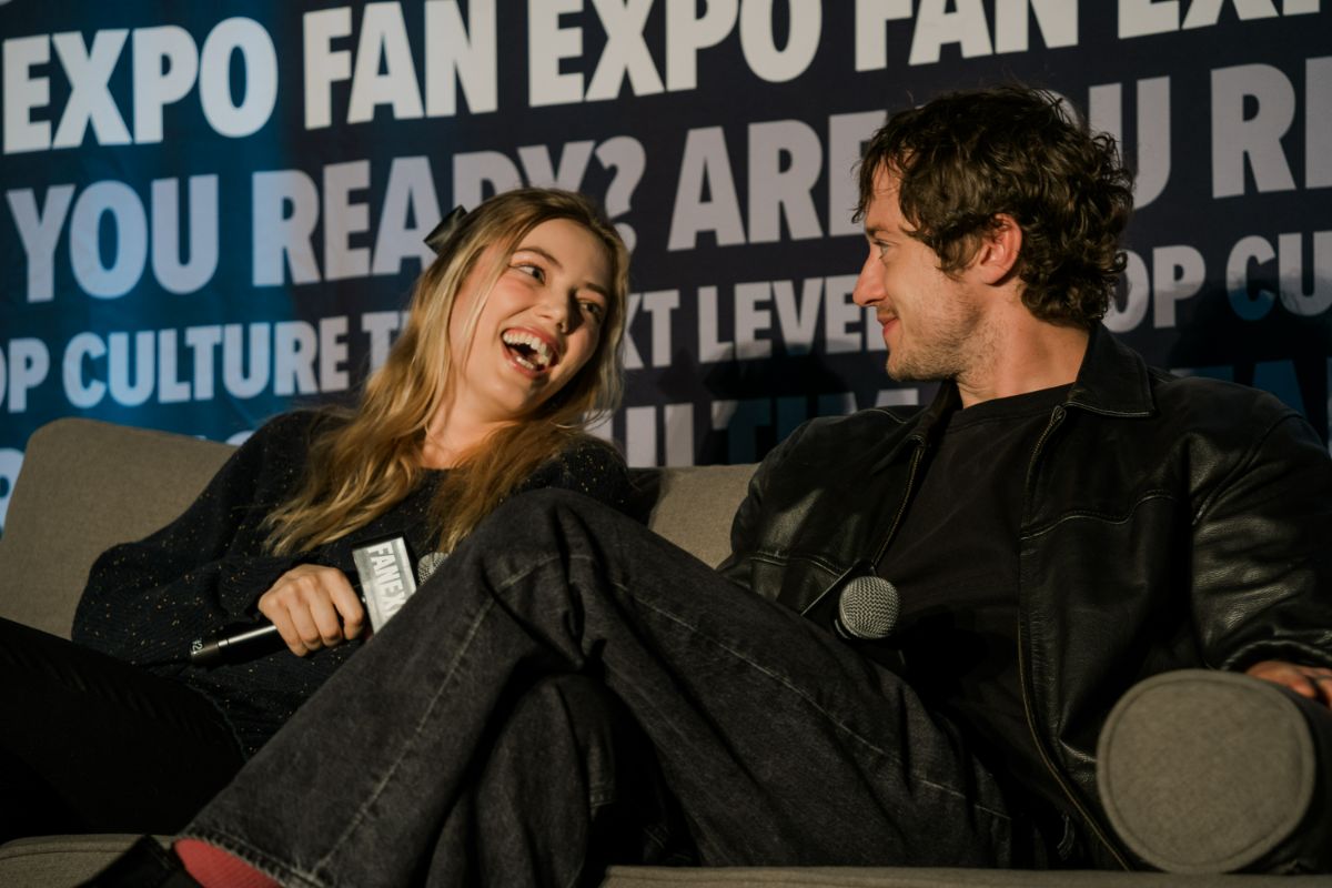 JOSEPH QUINN AND GRACE VAN DIEN SITTING ON A COUCH HOLDING MICROPHONES, LAUGHING AND SMILING