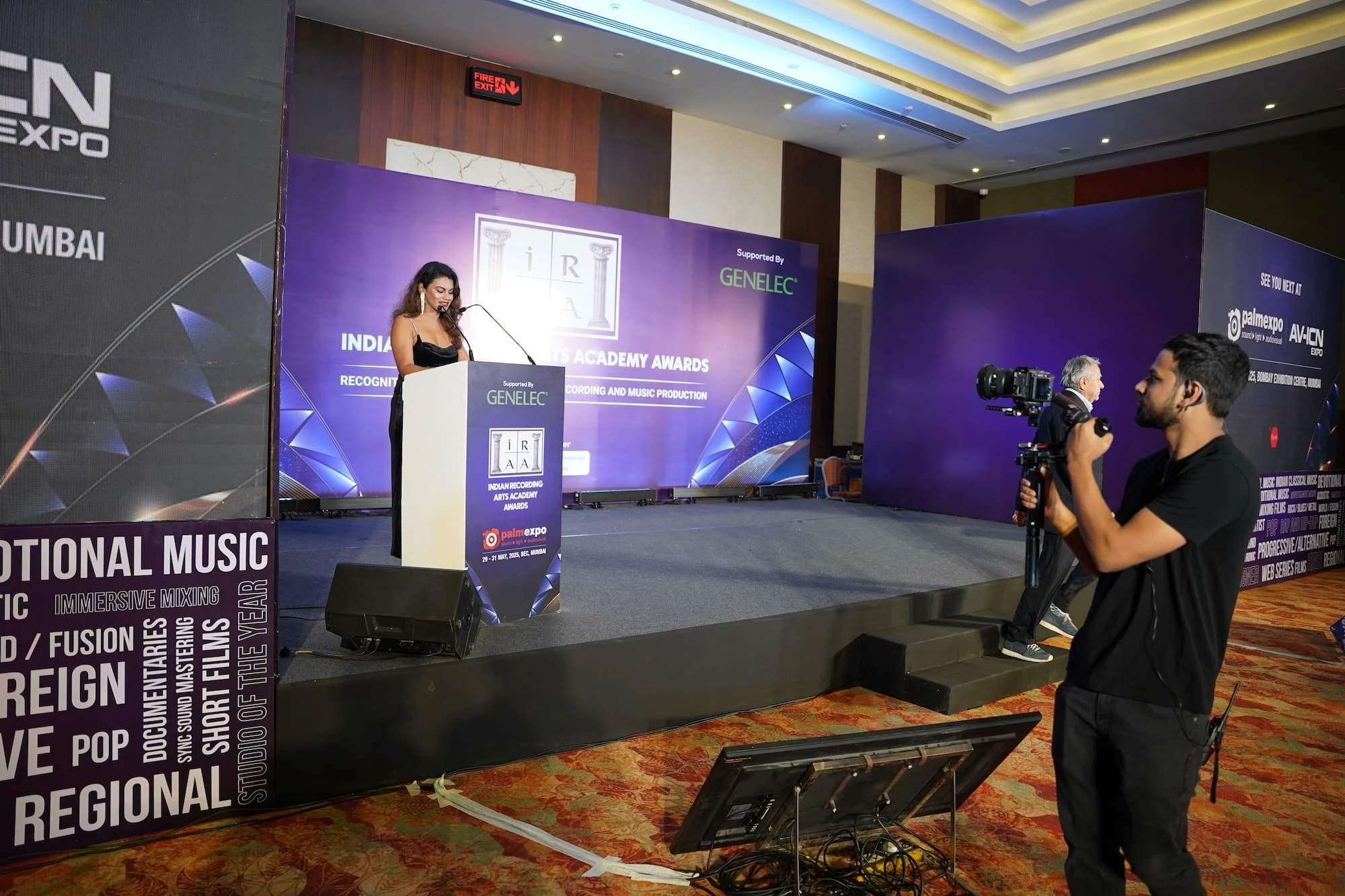 Woman speaking at a podium during an awards ceremony, with a cameraman filming the event at the IRAA Palm Awards