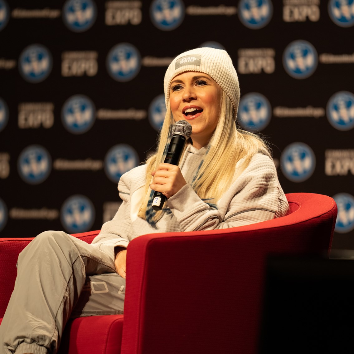 Star Wars legend Ashley Eckstein smiles while sitting on stage in a red chair & holding a microphone