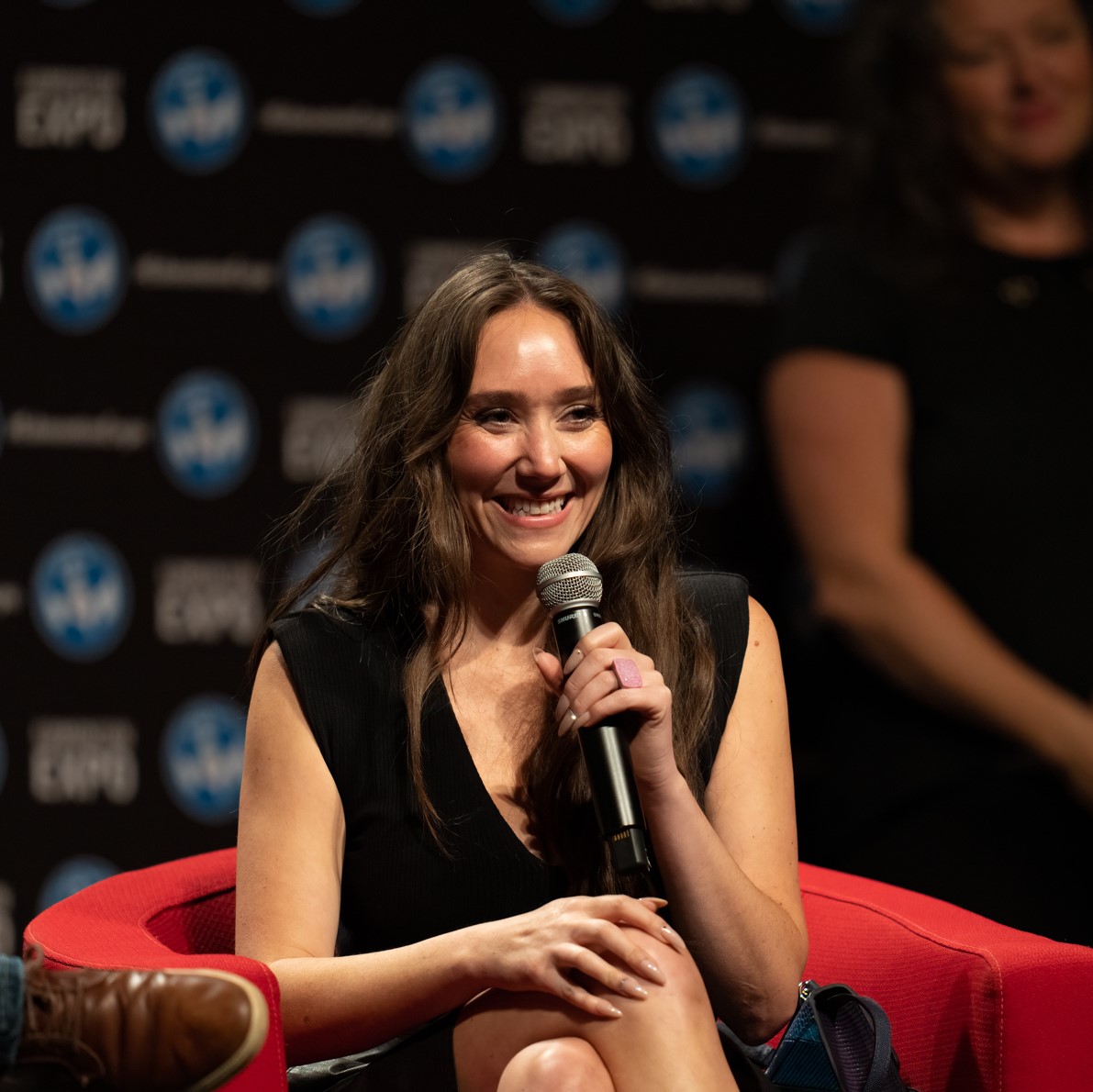 Patty Guggenheim is sitting in a red chair, holding a microphone and is smiling to the audience.