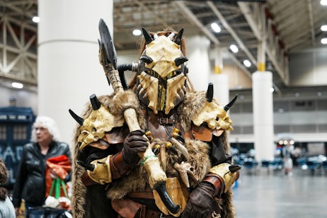 The image depicts an individual in an elaborate costume resembling a fantasy or alien creature. The costume includes a mask with horn-like protrusions and a design that mimics a skull, as well as armor crafted to look like it's made from bones. The wearer's pose, holding what appears to be a weapon, adds to the authenticity of the character's portrayal. This type of costume is typical of cosplay found at fan conventions, where attendees create and wear costumes of characters from various fictional universes, demonstrating a high level of detail and craftsmanship. The background suggests an indoor event space, likely a convention center.