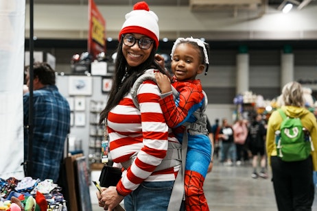 The image shows a smiling woman carrying a young child on her back. The woman is wearing glasses and a red and white striped hat and shirt, reminiscent of the popular "Where's Waldo?" character. The child is in a Spider-Man costume, complete with the iconic red and blue design. They both appear happy and are likely participating in a fun, themed event such as a comic book convention or a costume party. The environment around them is bustling, with various other attendees and booths in the background, indicative of a lively and family-friendly atmosphere at a public expo or similar gathering.