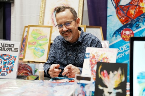 The image captures a cheerful man seated at a booth, surrounded by colorful artwork. He's wearing glasses, a blue patterned shirt, and is smiling warmly at the camera, giving the impression of a friendly and welcoming artist at his exhibit. In front of him and in the background, various pieces of vibrant art are displayed, including a framed picture that appears to feature cartoon-style turtles, which may suggest he is an illustrator or involved in character design. The booth setup with visible art pieces indicates this could be at an art fair, convention, or similar event where artists showcase their work. The man's engaging demeanor and the creative environment suggest he is likely interacting with attendees or fans.