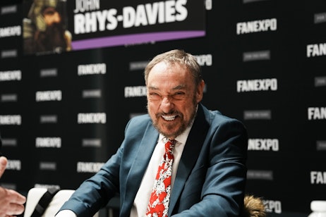 The image features John Rhys-Davies with a jubilant expression, seated and laughing, exuding a sense of joy and charisma. He is smartly dressed in a suit with a vibrant red tie that has a playful pattern, suggesting a personality that is both professional and lighthearted. The backdrop is adorned with the "FAN EXPO" logo, indicating that he may be a guest or speaker at a pop culture event. His engaging smile and the blurred background focus all attention on his animated interaction, capturing a moment of genuine happiness or amusement.