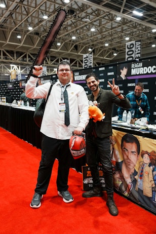The image captures Ryan Colt Levy with a male fan, both smiling and posing for the camera. The man on the left is dressed in a costume with a fake blood-splattered shirt, holding up a large prop weapon above his head. Ryan on the right is giving a peace sign, holding an orange plush toy and wearing a casual outfit with a jacket. Behind them is a backdrop with the name "REAGAN MURDOCK", indicating the setting is likely a signing booth or meet-and-greet area within the expo. Their cheerful expressions and playful poses reflect the fun and enthusiastic spirit of such events.