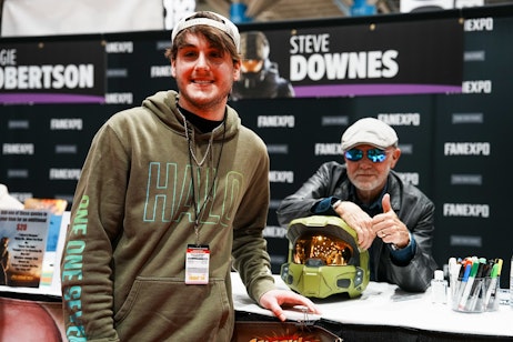 The image features two individuals at what appears to be a signing event. On the left is a young man smiling at the camera, wearing a cap and a hoodie with the word "HALO" printed on it, suggesting he is a fan of the video game series. He is wearing a lanyard with a badge, indicating his attendee status. On the right sits Voice Actor Steve Downes giving a thumbs-up sign, wearing reflective blue sunglasses and a black leather jacket, seated behind a table. In front of Steve is a helmet resembling that of the "Halo" game series protagonist, Master Chief, enhancing the thematic context of the event. A backdrop behind the table features the name "STEVE DOWNES." The event appears to be well-attended, as suggested by the presence of other people and the professional setup.