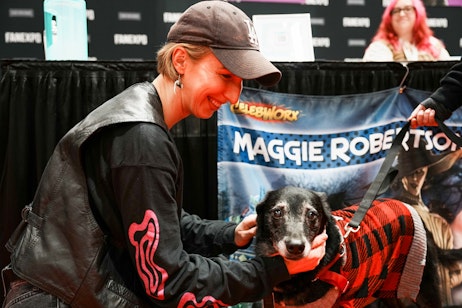 The image captures a heartwarming moment between voice actor Maggie Robertson and a dog at an event. She is leaning in with a joyful smile, gently petting the dog, which is looking directly at the camera with a relaxed expression. She is wearing a casual cap and a black leather jacket with a red design. In the background, a banner with the name "MAGGIE ROBERTS" suggests she may be at a signing or meet-and-greet area designated for someone of that name, possibly at a fan expo or convention given the "CELEBWORX" text also visible on the backdrop. The interaction between the woman and the dog adds a personal and tender touch to the setting.