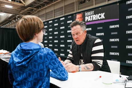 The image captures an encounter between a fan and Robert Patrick. The fan, whose back is to the camera, is wearing a blue patterned jacket and glasses, facing Robert across the table. Robert, with a prominent tattoo on his forearm, is wearing a black and white striped jacket and looks engaged in conversation with the fan. Behind the celebrity is a backdrop with his name, "ROBERT PATRICK," indicating his identity for those attending the event. The setting appears to be a convention hall, as indicated by the banner and the structure of the venue. The exchange seems friendly and personal, a common scene at fan expos where attendees can meet figures from popular culture.