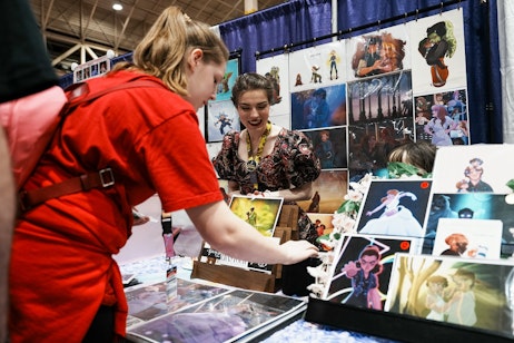 The image shows an interaction between two individuals at an artist's booth. The person on the left, wearing a red shirt and a crossbody bag, is browsing through artwork, seemingly selecting a piece. The artist, on the right, is smiling engagingly and watching the potential buyer with interest. She is wearing a dark floral dress and has a lanyard with a badge, indicating her participation in the event. The booth is adorned with a variety of colorful artworks displayed on the wall and table, suggesting a vibrant and creative setting. The atmosphere seems friendly and inviting, characteristic of an art fair or convention where artists showcase and sell their work.