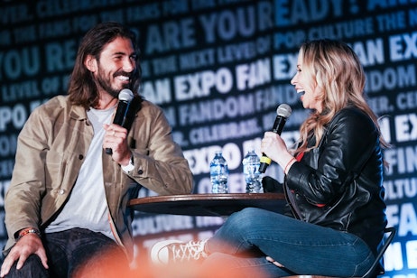 The image shows a man and voice actor Tara Strong sitting across from each other, both holding microphones and engaged in a conversation. They appear to be part of a panel or interview at a fan expo, as suggested by the words "FAN EXPO" visible in the background. The man, with long hair and a beard, wears a light jacket and a relaxed smile, while Tara, wearing a black leather jacket, is laughing. Their casual and engaged demeanors suggest a friendly and entertaining dialogue. Water bottles on the table indicate the event's duration, and the backdrop's text about the next level of pop culture hints at the expo's focus on entertainment media. Their comfortable posture and the candid capture of their expressions convey a moment of genuine interaction and enjoyment.