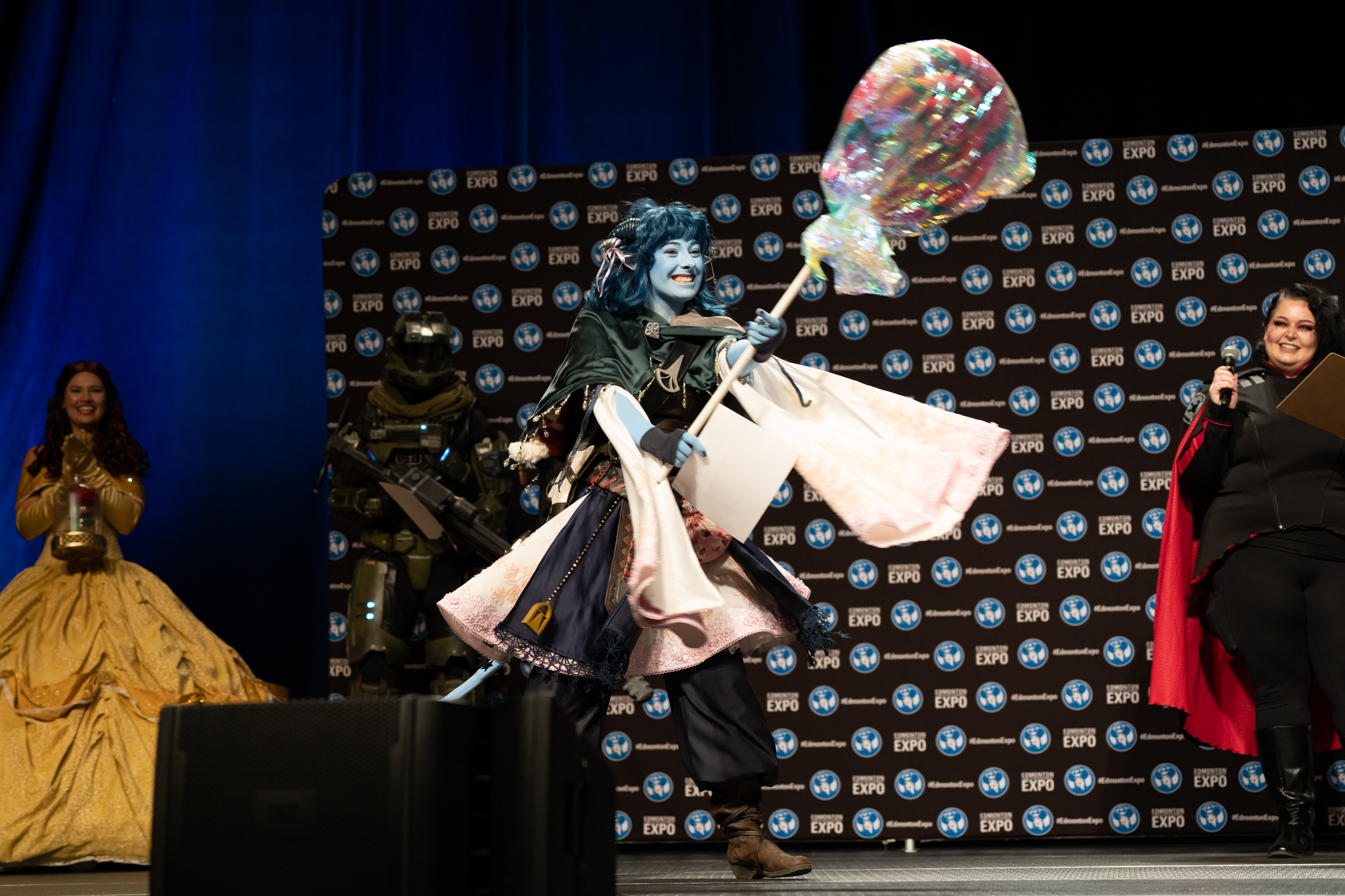 A cosplayer in blue face paint holds a giant lolipop prop as she smiles on stage
