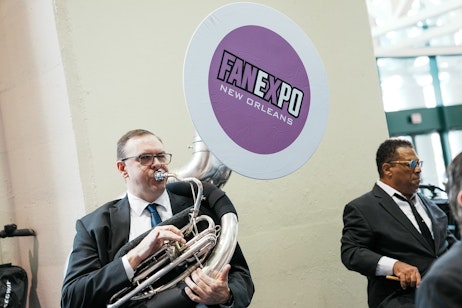 The image features a man playing a tuba, dressed in formal attire with a tie and glasses. The tuba has a circular cover with the logo "FAN EXPO New Orleans," indicating that he is performing at a fan convention in New Orleans. The musician's focused expression while playing the instrument suggests he is professionally contributing to the event's atmosphere, providing live music for the attendees. The simple background with neutral colors keeps the attention on the performer and the FAN EXPO branding.