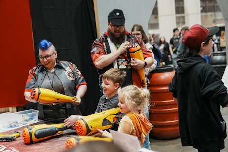 The image shows two adults and two children at an event, engaging with large toy blasters. The adult on the left, with striking blue hair, is watching the children with a delighted expression, sharing in their excitement. The man on the right, with a beard and wearing a cap, is helping, or supervising. Both children are focused on the blasters, with the boy in the center holding one and the girl on the right reaching out to touch it, both appearing thrilled with the experience. The background has a simple black curtain, which suggests an indoor venue, possibly a toy convention or a recreational event. The atmosphere is lively and filled with joy, capturing a moment of playful discovery and interaction.