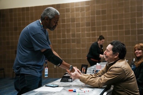 The image captures a moment of warm interaction between Peter Cullen and a fan at an indoor event. The man on the left, standing, extends his hand towards Peter on the right, who is reaching out to reciprocate the handshake. Both men appear to be engaging in a friendly and respectful greeting or acknowledgment. Peter is dressed in a casual jacket and seems to be at a signing table, as indicated by the markers and items in front of him. The environment suggests a convention or meet-and-greet setting, with the tiled wall in the background adding to the indoor atmosphere. The exchange between the two men is the focal point, illustrating a personal connection often found at such events.