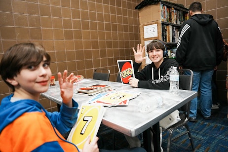 The image shows two young people playing a giant version of the card game UNO. The child in the foreground is holding a large yellow card with the number 7 on it, while the other child is waving with a smile and holding up a card that has the UNO game logo. They are seated at a metallic table with more oversized UNO cards spread out in front of them. The setting appears to be an indoor public area, possibly a recreational room or a school setting, as indicated by the brown-tiled walls and the bookshelf in the background. Both children are casually dressed, with the one in the foreground wearing a blue and orange jacket, and the one in the background wearing a black hoodie. Their expressions and gestures suggest they are having a fun and engaging time.