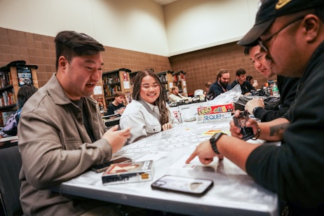 The image shows a man and a woman seated at a table, both with joyful expressions, engaged in a board game. The man is on the left, wearing a grey shirt, and appears to be in the midst of a lighthearted conversation or action, gesturing with his hand. The woman on the right is smiling broadly, dressed in a light-colored coat. On the table between them are cards laid out in an orderly fashion, part of the game they are playing. There is a smartphone and a boxed board game titled "SEQUENCE" visible on the table, indicating the type of game they might be playing. The setting looks like a public indoor space, possibly a board game cafe or a convention, with other people in the background also seated and engaged in similar activities.