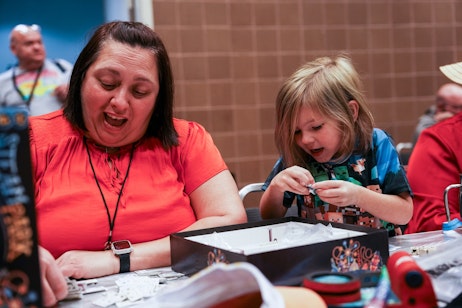 The image shows a woman and a young boy engaged in an activity together, with the woman looking on with an expression of excitement. The woman is wearing a bright orange top and has a watch on her left wrist, and her hair is styled in a short, straight manner. The boy is focused on something in his hands, possibly a game, with a look of concentration. He is wearing a blue shirt with a pixelated design. They are sitting at a table, and there are small pieces, which could be part of a game or craft, scattered in front of them. The background suggests they are indoors, possibly in a public place like a school or community center, with a tiled wall visible behind them.
