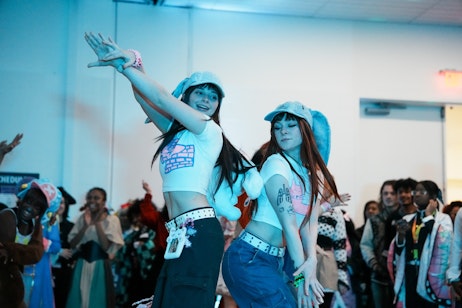 The image captures two individuals mid-dance, with one extending her arm skyward and the other with her arm swept to the side. They both wear cropped white tops, casual pants with decorative belts, and matching bucket hats, giving off a coordinated, energetic vibe typical of a dance or performance troupe. Their expressions are joyful and focused, typical of performers enjoying their routine. The scene is likely part of a larger event, possibly a dance competition or exhibition, suggested by the audience in the background and the informal setting.