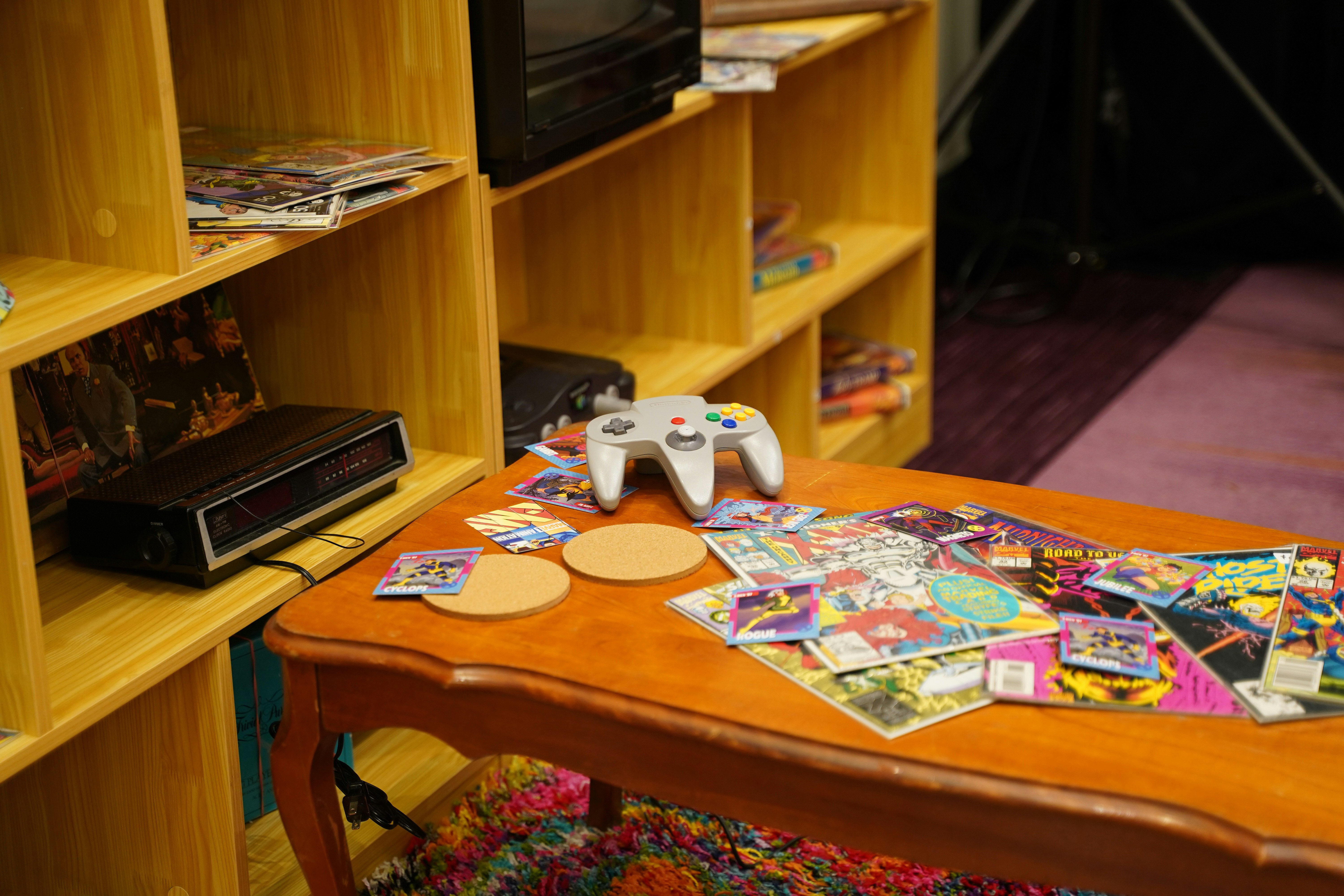 A COFFEE TABLE COVERED IN OLD COMICS, AND TRADING CARDS, SITTING ON THE CORNER IS A SUPER NINTENDO CONTROLLER. NEXT TO THE TABLE, A LARGE SHELF HAS AN ALARM CLOCK, ACTION FIGURES, AN OLD BOX TV, COMICS, AND VHS TAPES.