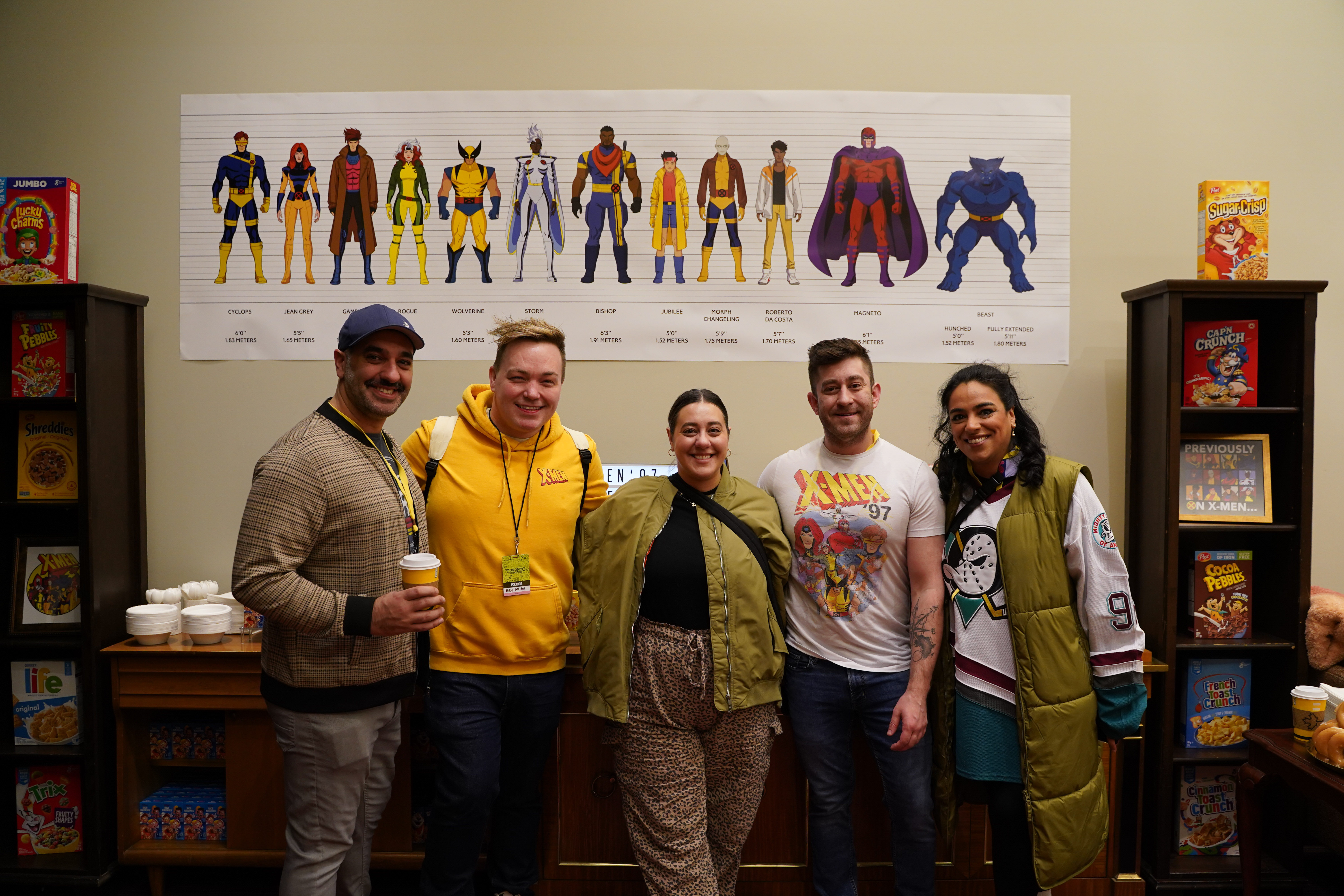 Fans gather at the breakfast bar for a photo in Xavier's Lounge for Extraordinary Youngsters, behind them is a police line up style image of the X-Men