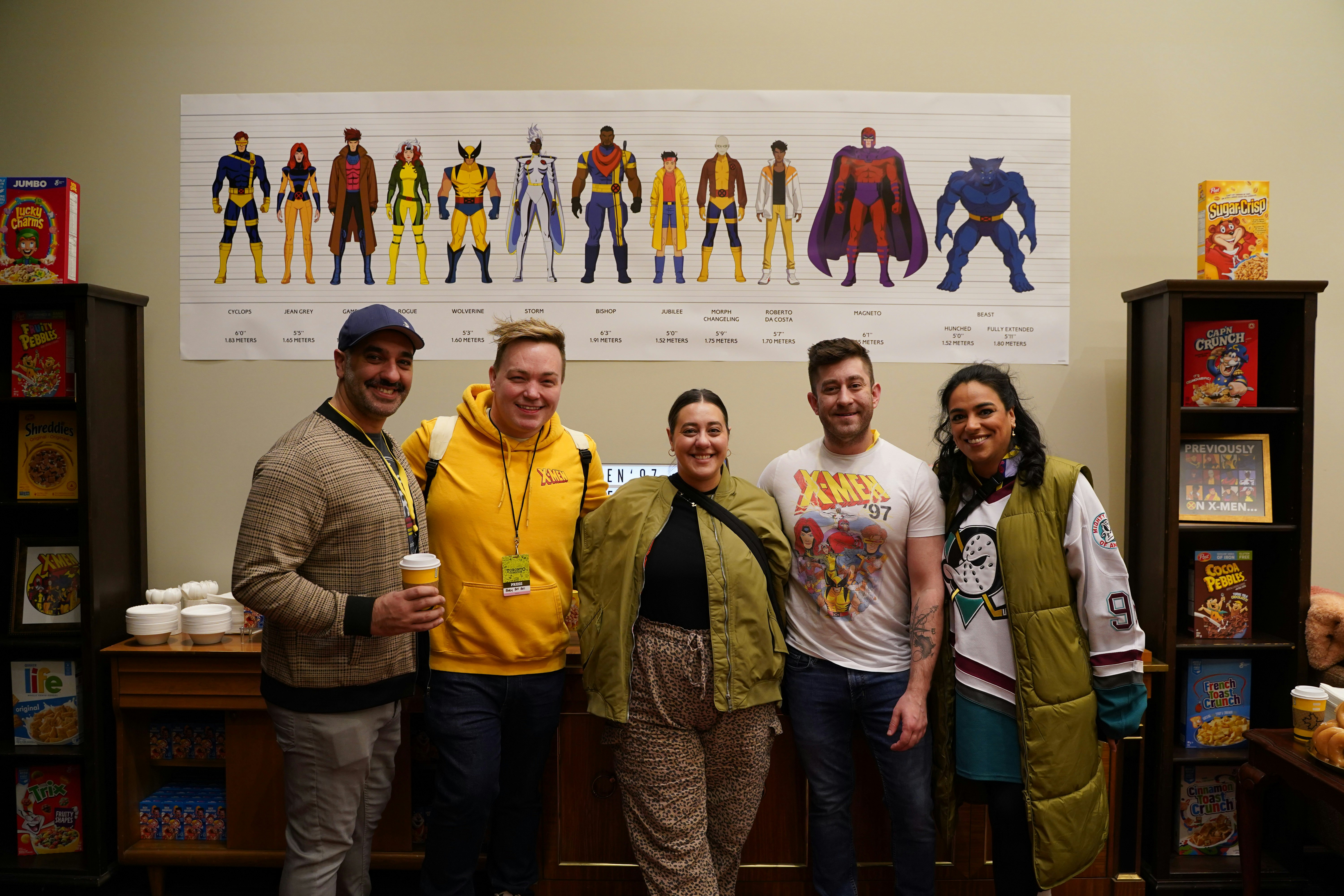 Fans gather at the breakfast bar for a photo in Xavier's Lounge for Extraordinary Youngsters, behind them is a police line up style image of the X-Men