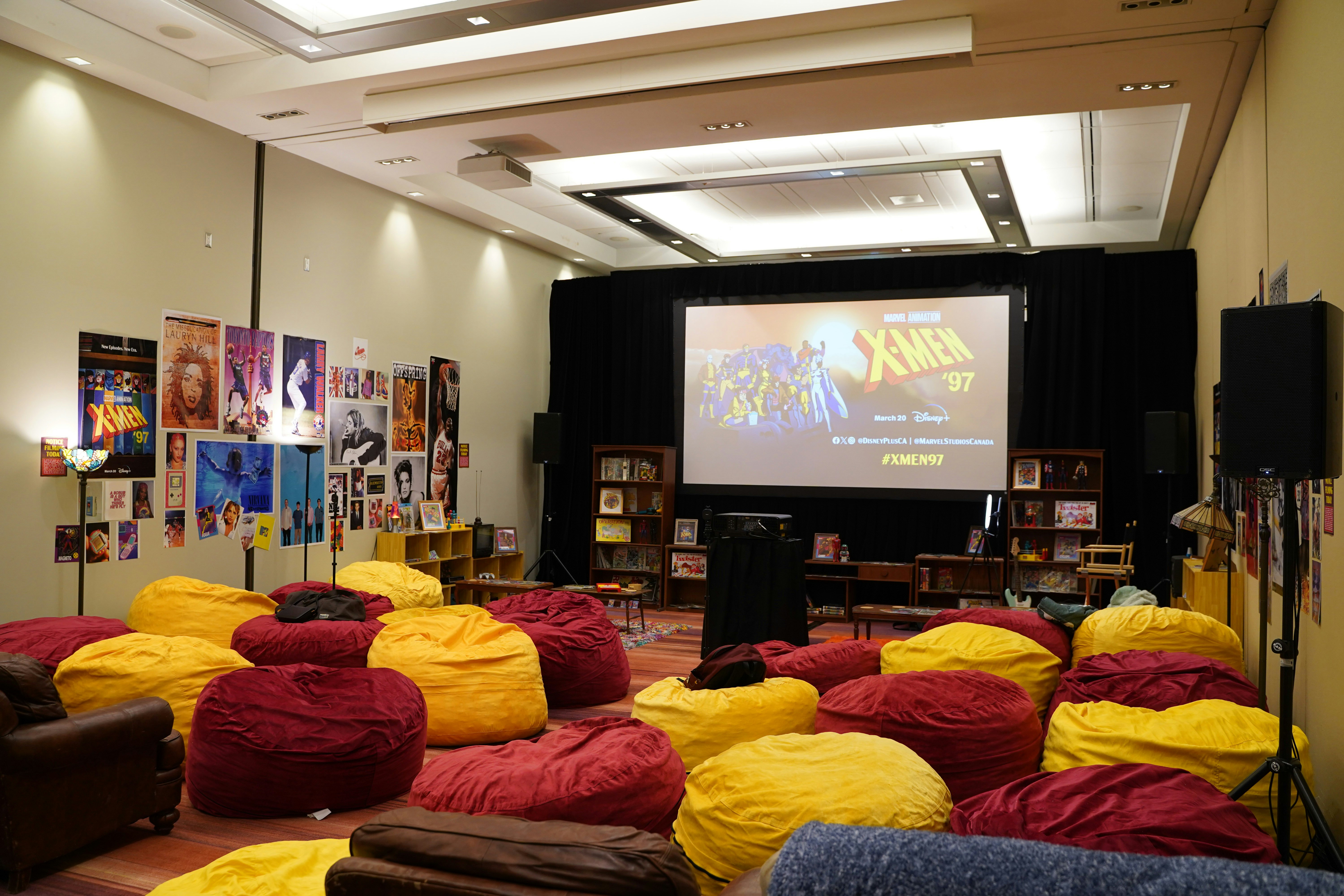 A wide shot of Xavier's Lounge before fans enter. the room is full of red and yellow bean bag chairs. The walls are covered in posters featuring 90s music artists, television shows, and movies. On the back wall, a screen, being illuminated by a projector showing the X-Men '97 promo art.