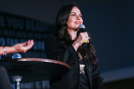 The image shows Lana Parrilla speaking into a microphone, looking upward with a thoughtful and pleasant expression. She is dressed in a smart black blazer with a sequined detail, suggesting a formal or professional setting. Her engagement with the audience, indicated by her open posture and attentive look, implies she might be answering a question or delivering a speech. The darkened background focuses the viewer's attention on her, indicating that she is the main speaker at the event, possibly a panel or a presentation at a conference or convention.
