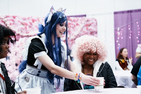 The image captures a joyful moment between two people. On the left, a person is dressed in a maid-style costume with blue hair, cat ears, and a tiara, indicative of cosplay. They are leaning forward with a bright, playful smile. On the right, a person is laughing heartily, sporting a curly pink wig and colorful bracelets, also suggesting a festive or cosplay-related event. The background is soft and blurred with pink hues, which may be part of a themed setup or decoration. The two individuals seem to be sharing a lighthearted and happy interaction, typical of the friendly and enthusiastic atmosphere found at conventions and cosplay events.
